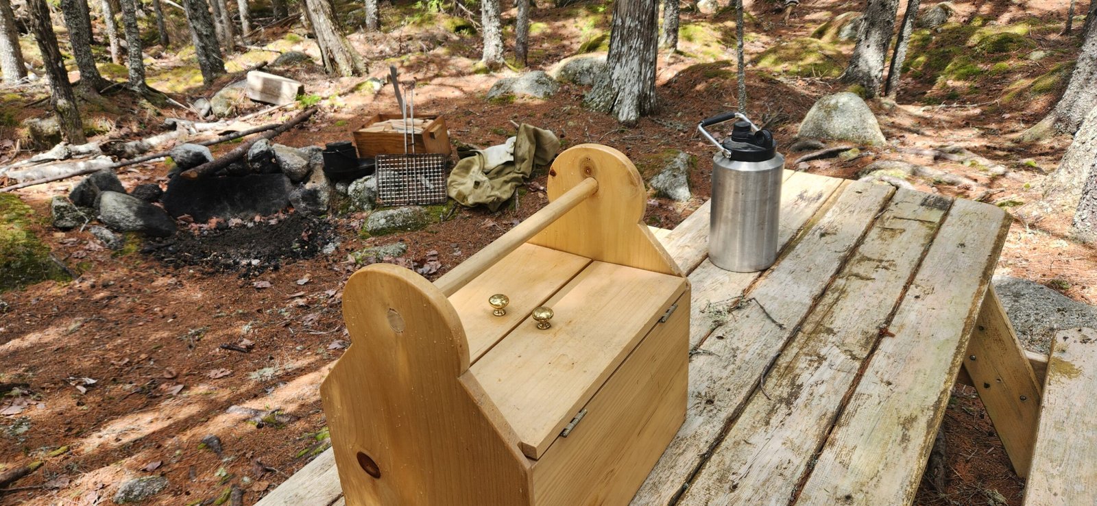 Rustic campfire setup with wooden gear box and cooking tools in a Maine forest