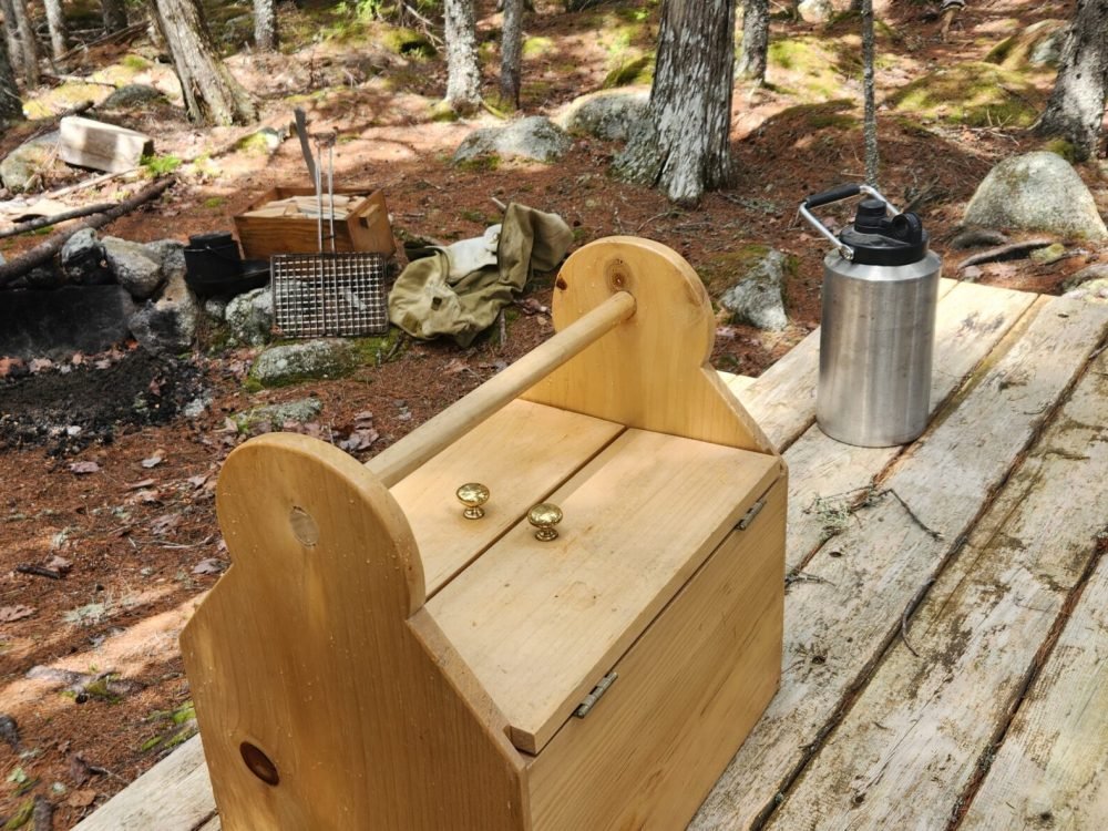 Rustic campfire setup with wooden gear box and cooking tools in a Maine forest